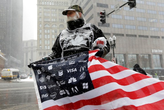 Protester Bob Bowes, of Somerville, Mass., displays an American flag featuring corporate logos, outside the location of a news conference by General Electric CEO Jeff Immelt, Massachusetts Gov. Charlie Baker, and Boston Mayor Marty Walsh, Monday, April 4, 2016, in Boston. Bowes joined a protest with others outside the news conference to highlight the millions of dollars in tax breaks and public incentives used to lure the company. (AP Photo/Steven Senne)